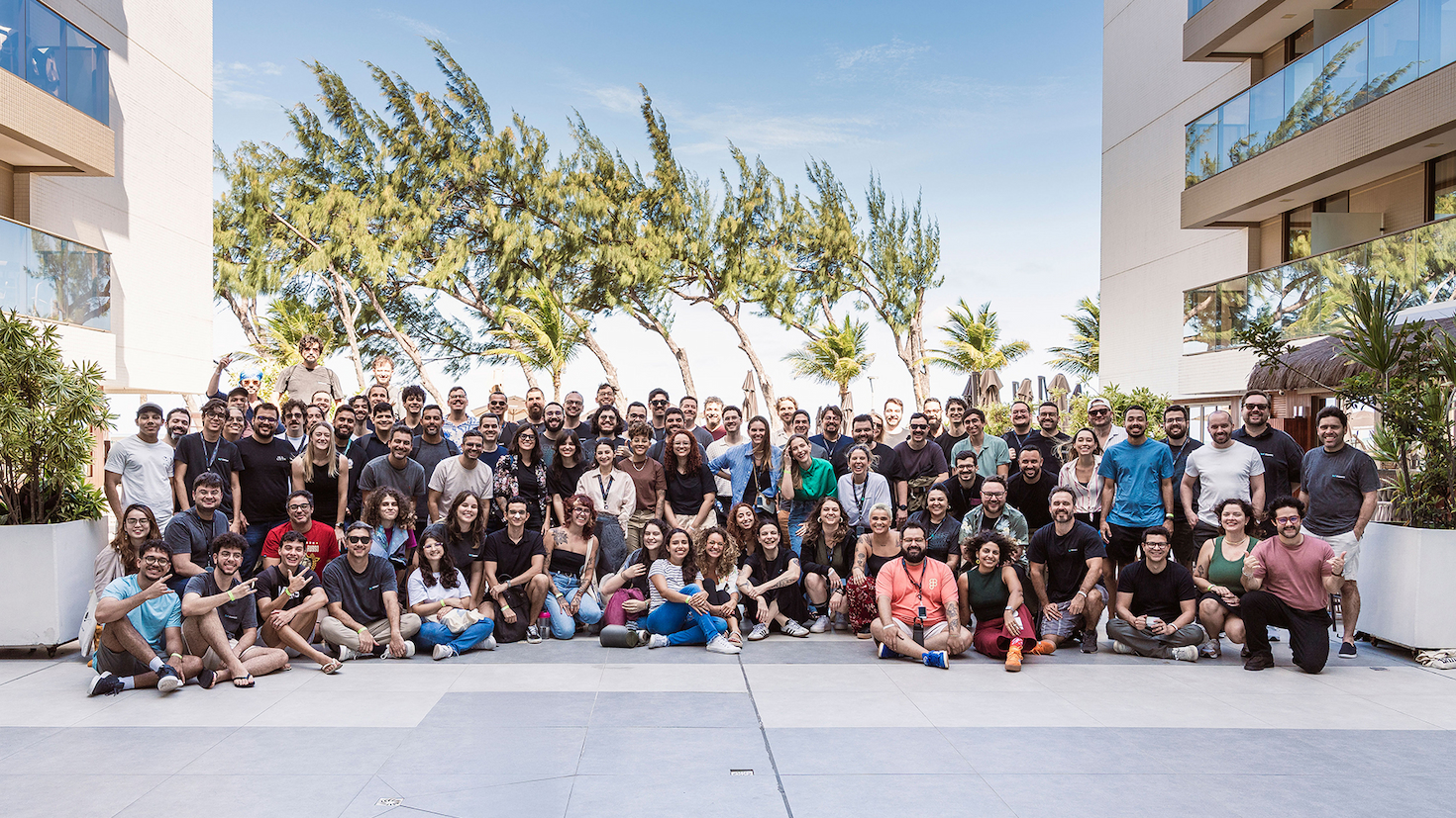 A group photo of the Moises team, gathered outside in a plaza surrounded by large potted plants, glass-rimmed balconies, and trees swaying in the distance.