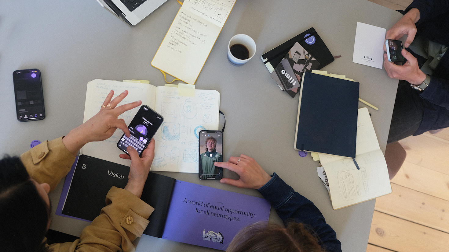 A top-down photo of people at a desk. On the desk are notebooks, iPhones, a coffee, and scattered items for note-taking and organization.